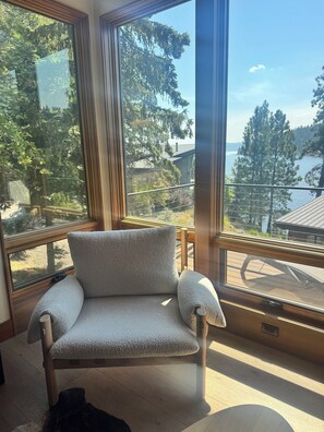 Una habitación con un gran ventanal, un sillón gris y vistas a un bosque y al agua.