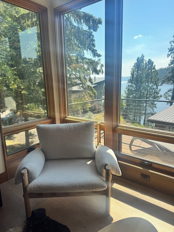 A room with a large window, a grey armchair, and a view of a forest and water.