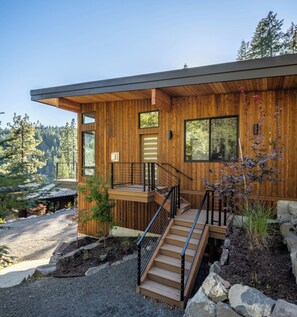 A modern wooden cabin with a deck, a staircase, and a view of a forest.