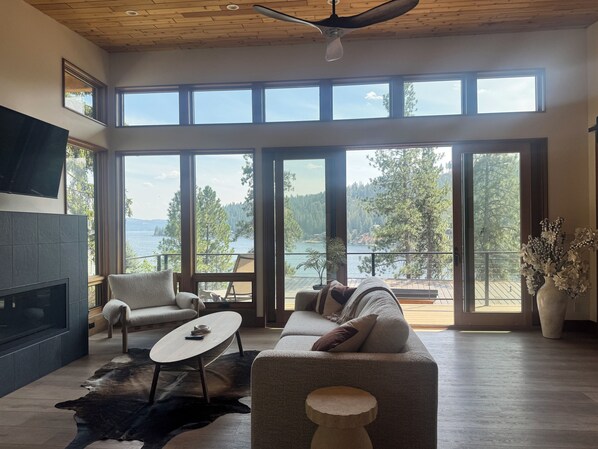 Amplia sala de estar con chimenea, asientos cómodos y ventanas grandes que ofrecen vistas a un lago y un bosque.