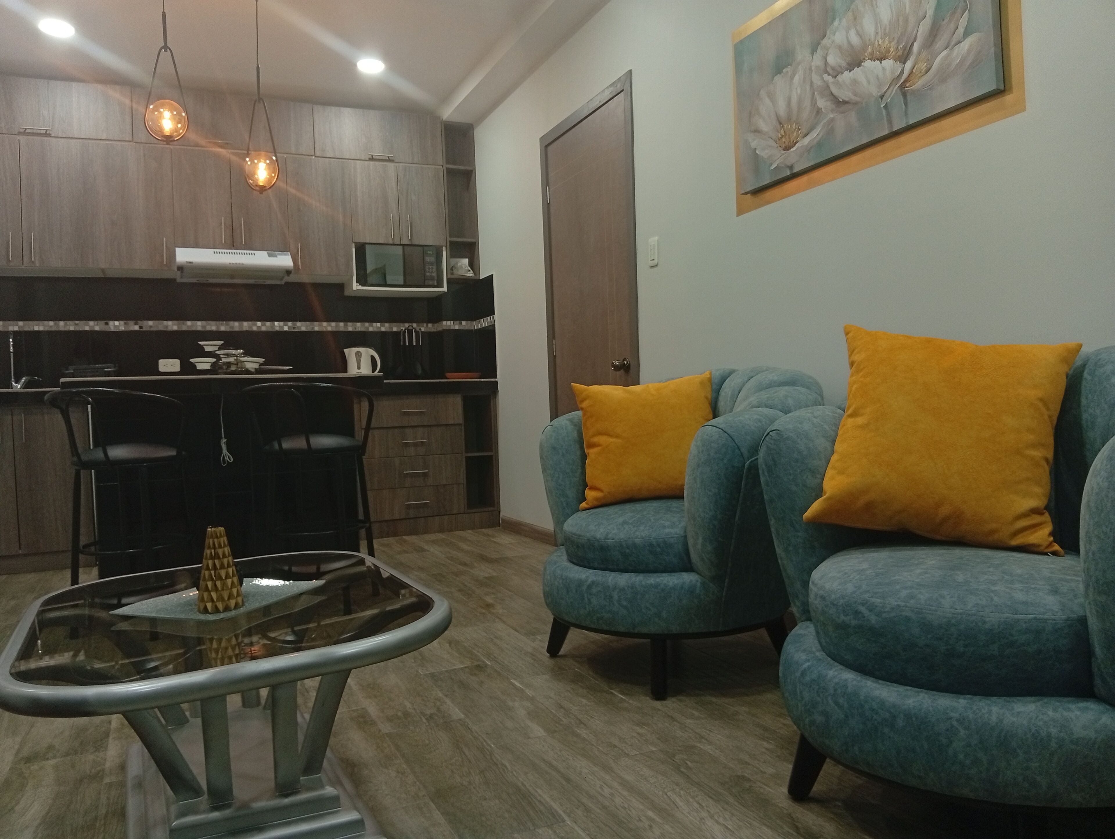 Ein modernes Wohnzimmer mit einem Glastisch, einer Couch mit orangefarbenen Kissen und einer Küchenzeile mit Holzschränken und Pendelleuchten.