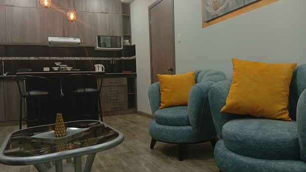 Un salon moderne avec une table basse en verre, un canapé agrémenté de coussins orange et un coin cuisine doté de meubles en bois et d’un éclairage suspendu.