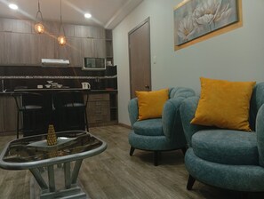 Una sala de estar moderna con una mesa de centro de vidrio, un sofá con cojines naranjas y una zona de cocina con armarios de madera e iluminación colgante.