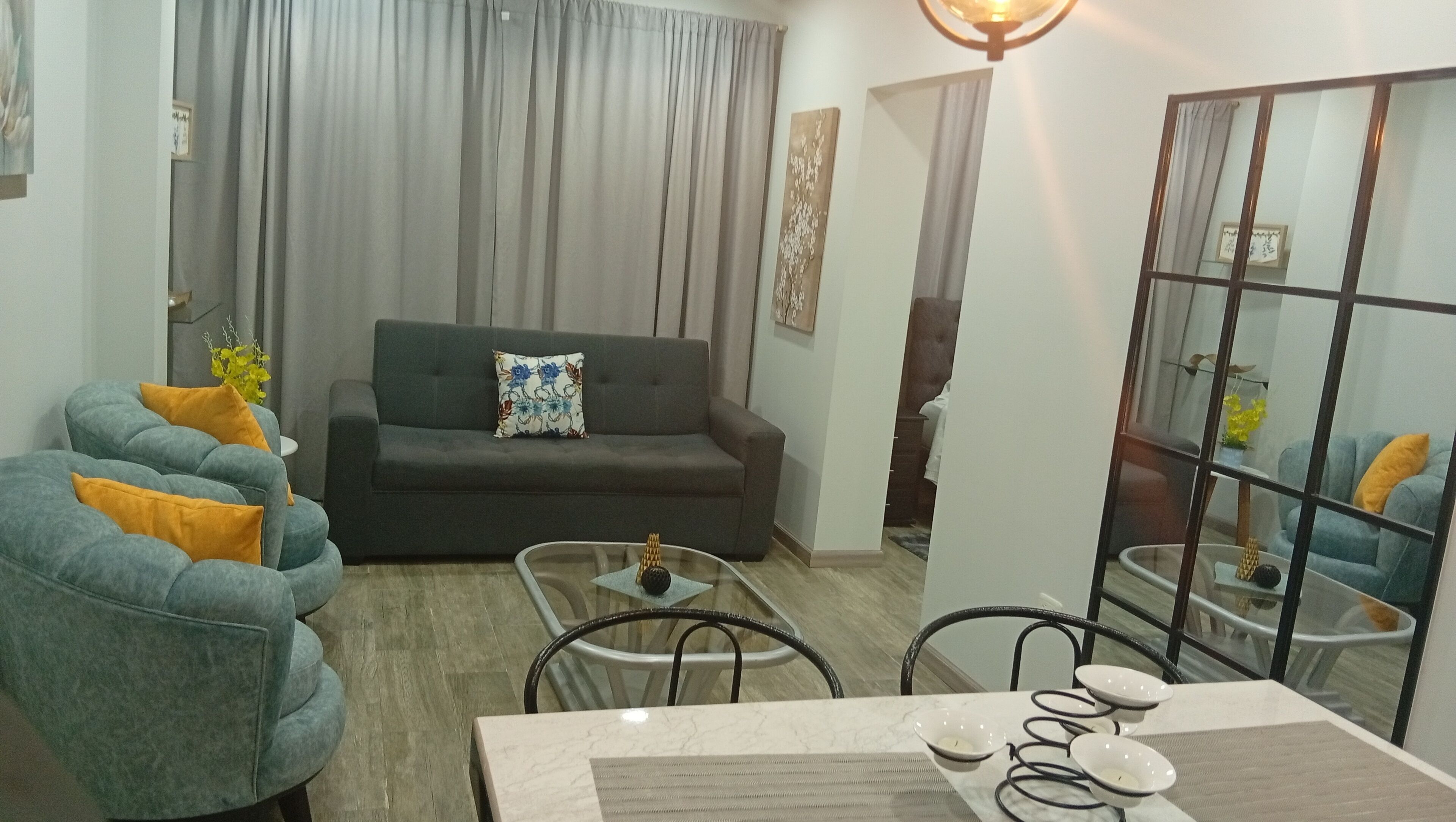 A living room with a sofa, a coffee table, and a dining table with chairs. There is a large window with curtains, a wall-mounted light fixture, and a mirror reflecting part of the room.
