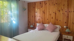A bedroom with a wooden paneled wall, a bed with white bedding and pink pillows, two bedside tables with lamps, and a hanging plant. - Casa Pil Riogordo by Ruralidays (Málaga)
