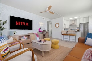 A modern living room with a flat-screen TV, a sofa, a coffee table, and a kitchen area with white cabinets and a stainless steel refrigerator.