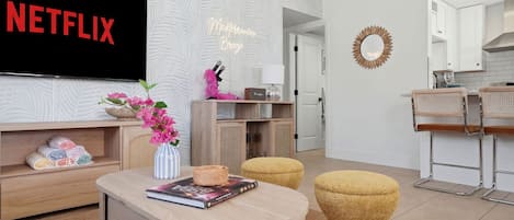 A modern living room with a flat-screen TV, a bookshelf, a coffee table, and a kitchen area in the background.