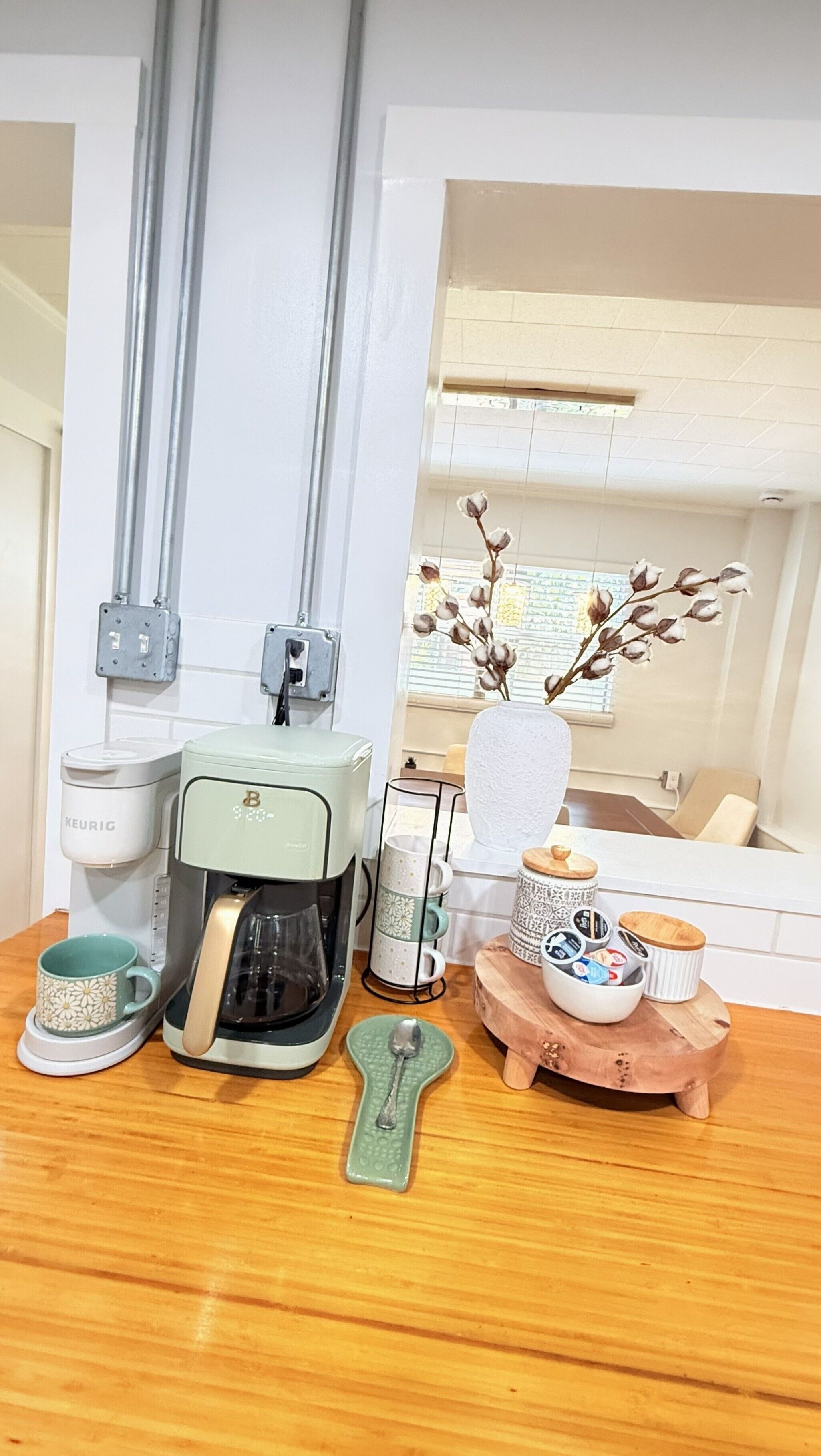 A kitchenette with a coffee maker, a kettle, a mug, a spoon, and a vase with flowers.