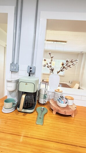 Una kitchenette con cafetera, tetera, taza, cuchara y un florero con flores.