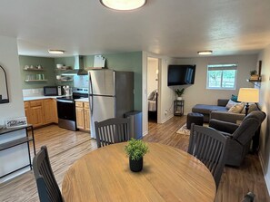 A modern kitchen with stainless steel appliances, a dining area with a round table, and a living room with a sofa and TV.