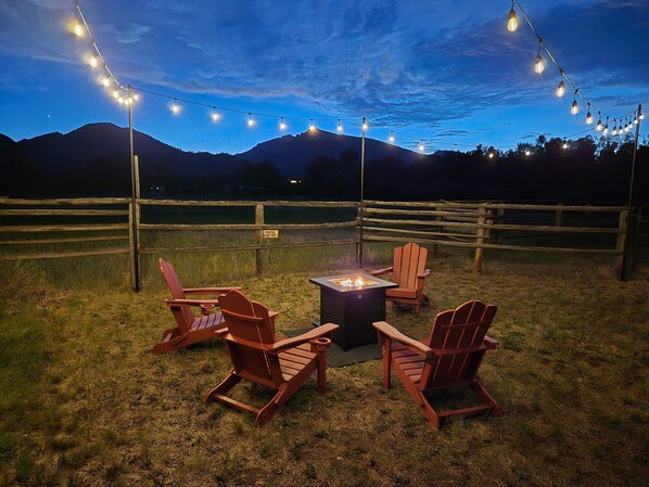 An outdoor seating area with Adirondack chairs and a central fire pit, surrounded by a wooden fence and string lights.