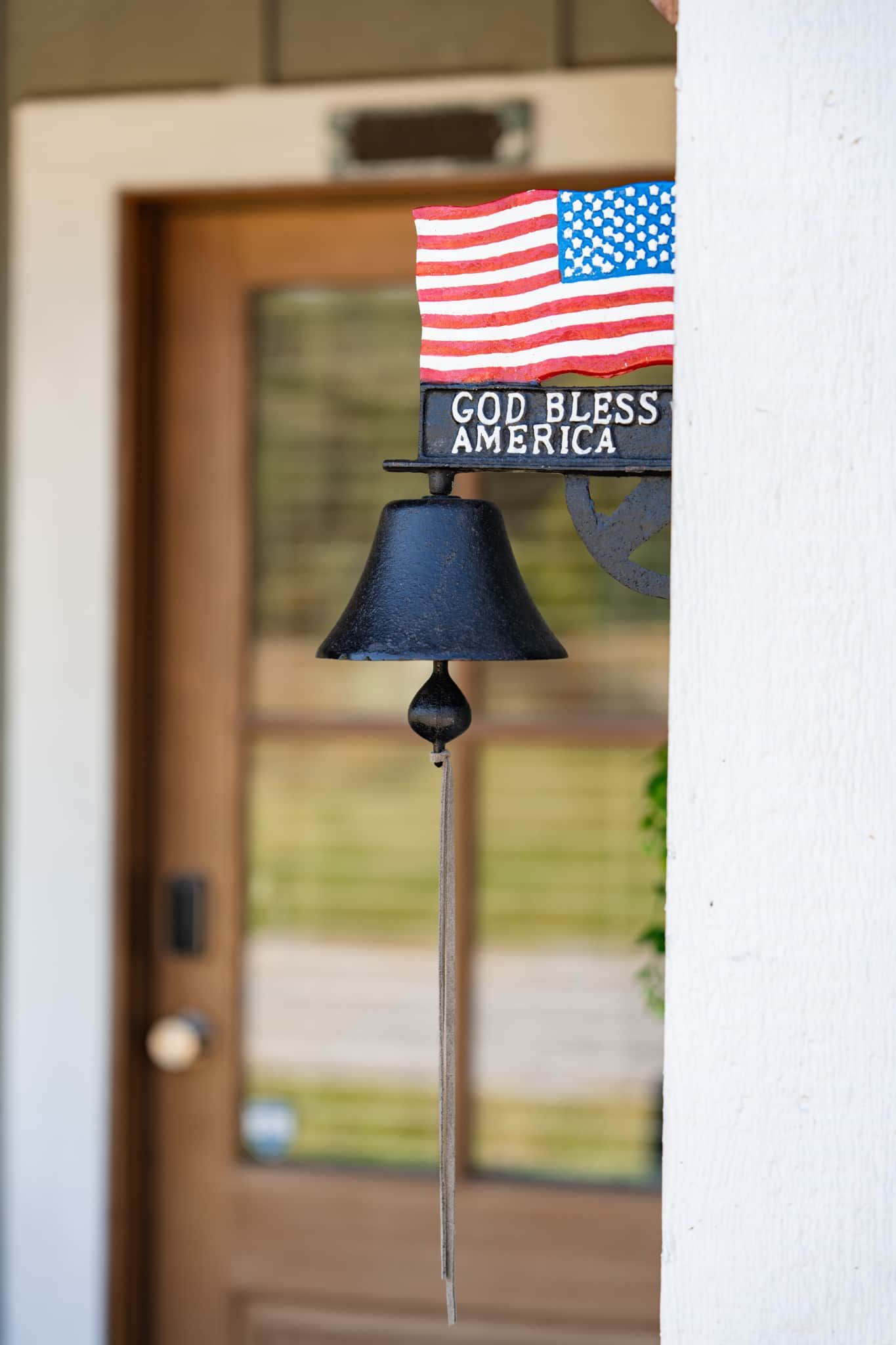 Una campana in metallo nero con una bandiera e un cartello con la scritta 'GOD BLESS AMERICA' montata su una parete bianca.