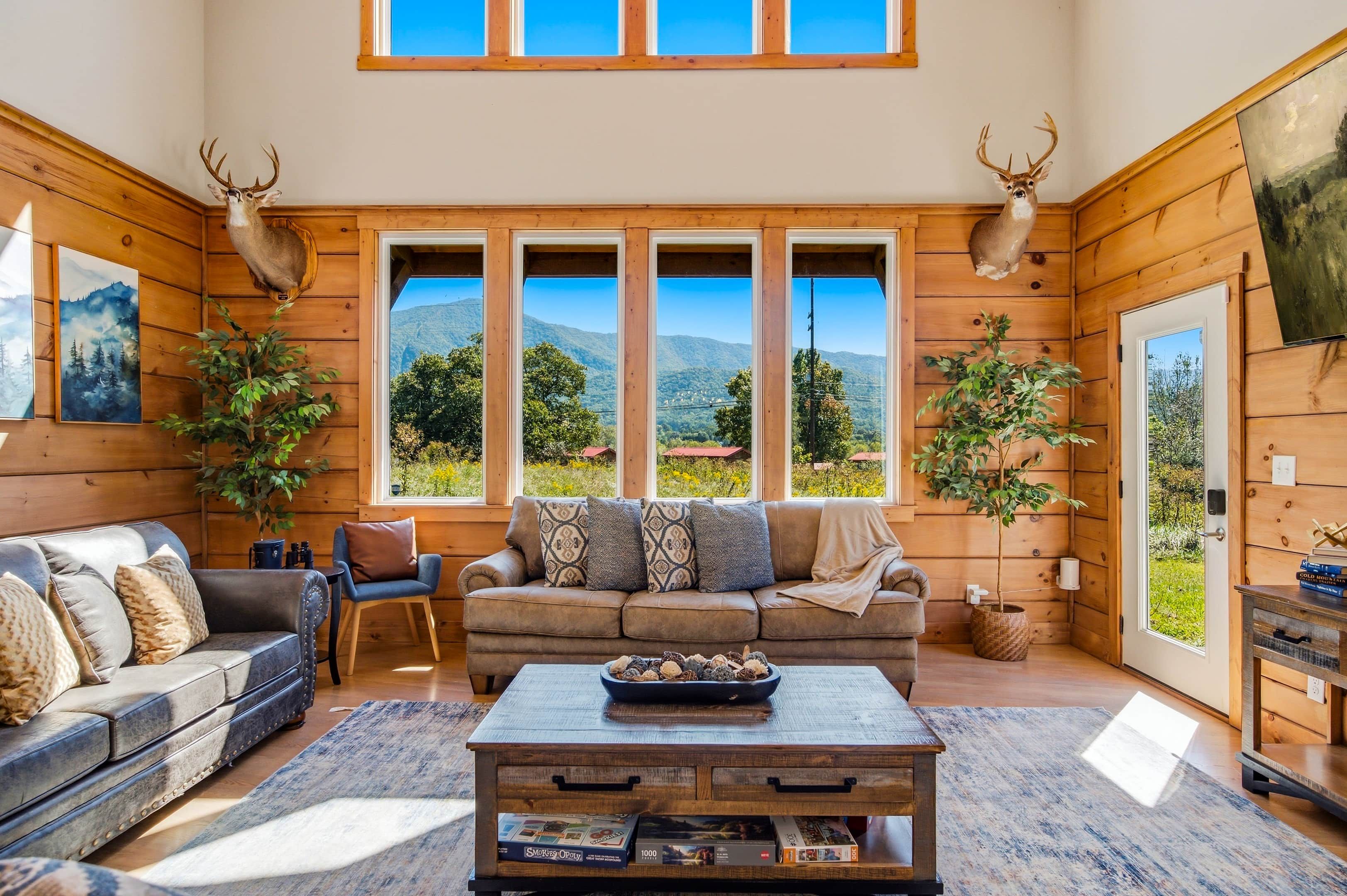A cozy living room with a large window, wooden walls, a sofa, a coffee table, and decorative antlers.