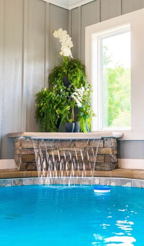 Uma piscina com cascata, rodeada por plantas em vasos e uma janela.