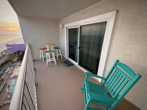 Ein Balkon mit Blick auf einen Strand, eine Glastür, ein türkisfarbener Stuhl und ein kleiner Tisch mit Hockern.