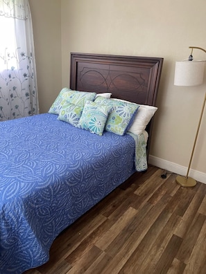 A bed with a blue patterned bedspread, multiple pillows with paisley designs, a wooden headboard, a floor lamp, and a window with a floral curtain.