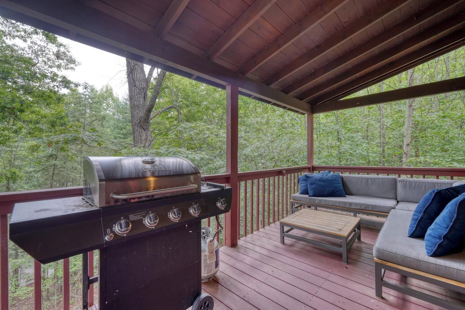 A covered outdoor deck with a grill, a sofa with cushions, and a wooden table.