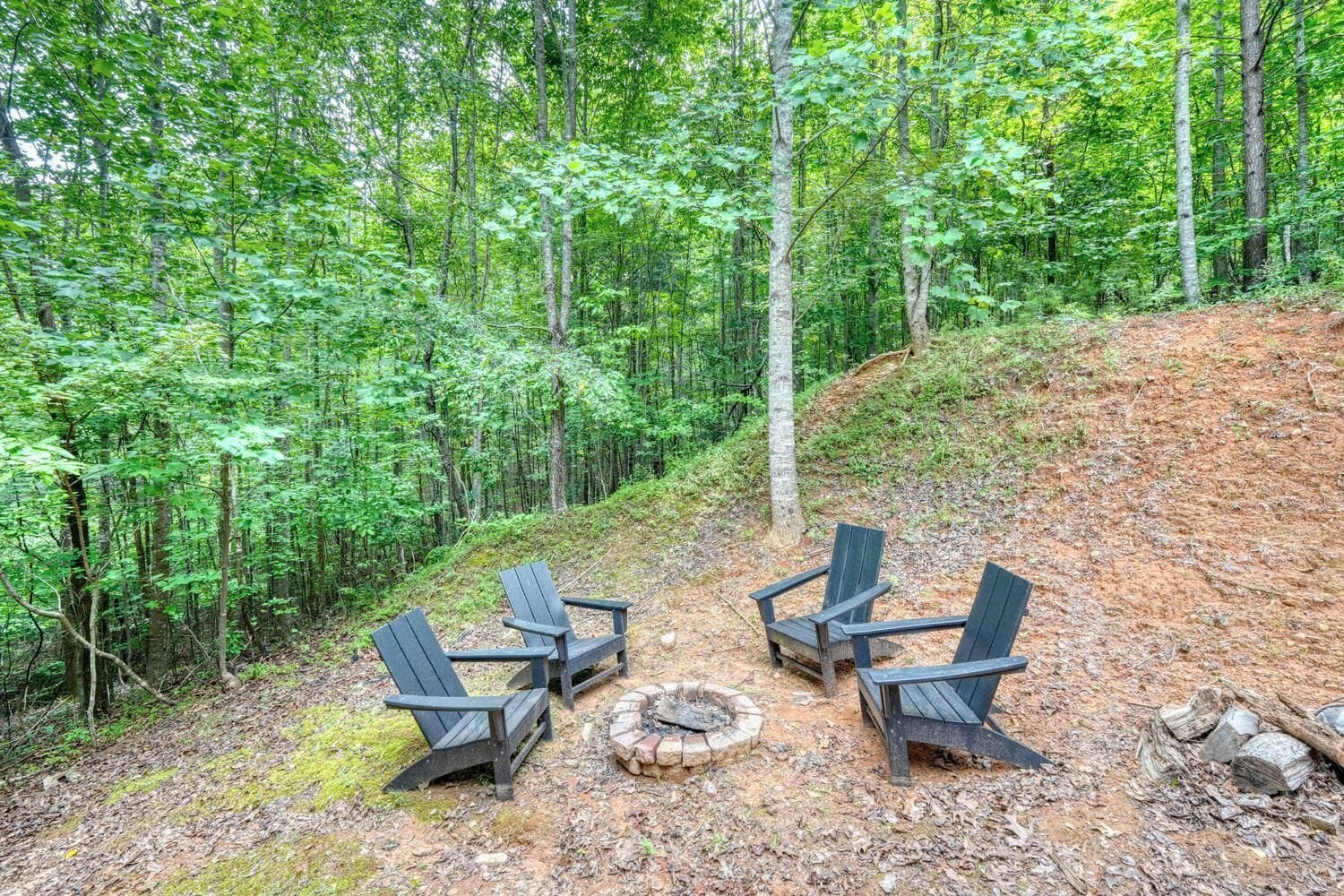 A forested area with a fire pit and three Adirondack chairs.