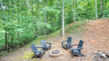 A forested area with a fire pit and three Adirondack chairs.