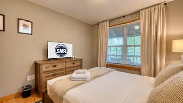 Quarto de hotel com uma cama, uma televisão sobre uma cômoda de madeira, uma janela com cortinas e uma luminária fixada na parede.