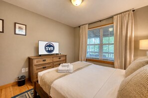 Quarto de hotel com uma cama, uma televisão sobre uma cômoda de madeira, uma janela com cortinas e uma luminária fixada na parede.