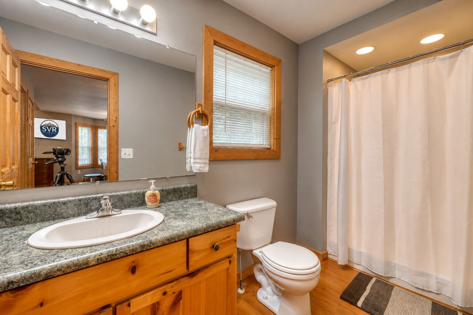 A bathroom with a double sink vanity, a toilet, a shower curtain, and a mirror with a logo.