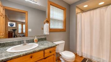 A bathroom with a double sink vanity, a toilet, a shower curtain, and a mirror with a logo.