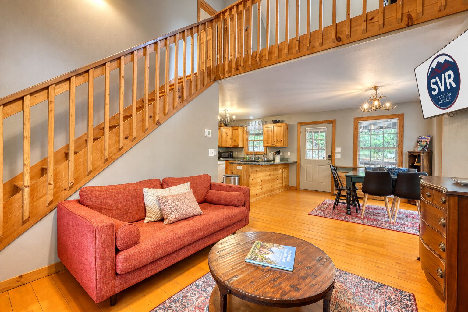 A spacious living area with a red sofa, wooden staircase, kitchen, dining area, and a sign for vacation rentals.