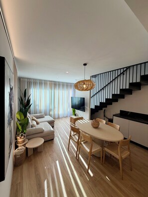 Een moderne woonkamer met een eethoek, een trap en een groot raam met zonwering.