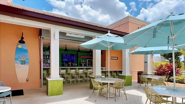 Ein Terrassenbereich im Freien mit runden Tischen und Stühlen, Sonnenschirmen und einer Bar mit einem Fernsehbildschirm.