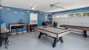 Ein Freizeitraum mit Billardtisch, zwei weiteren Billardtischen, Deckenventilatoren, Lichterketten und einem Fenster mit Jalousien.