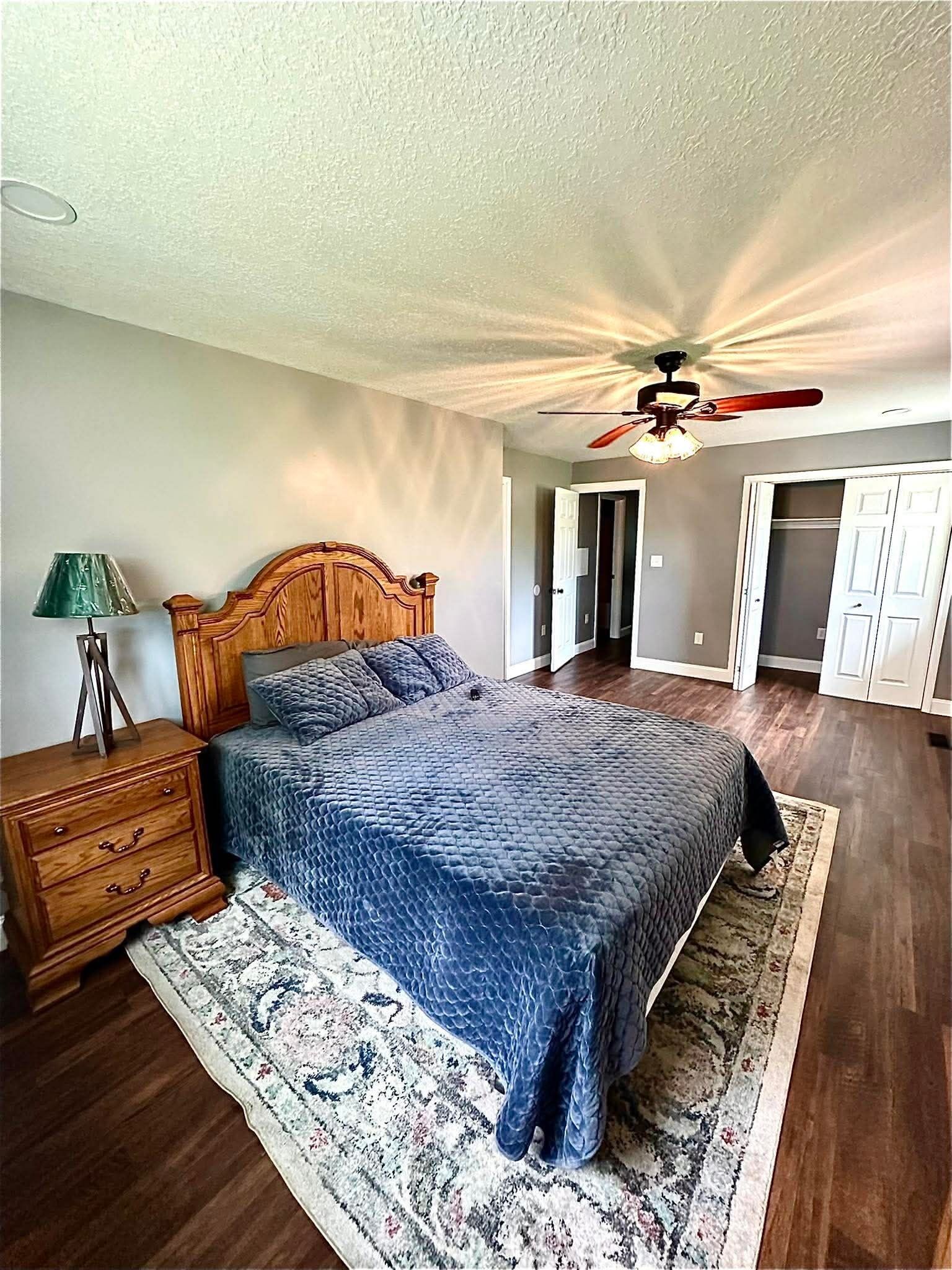 A bedroom with a wooden bed, a nightstand, a lamp, a ceiling fan, and a patterned rug.