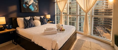 Un dormitorio moderno de hotel con una cama grande, vistas a la ciudad y un cuadro en la pared.