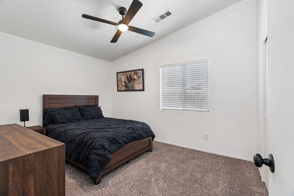 A bedroom with a bed, a ceiling fan, a window with blinds, a wall-mounted picture, and a door.