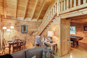 A wooden interior with a dining area, a sofa, a staircase, and a pool table.