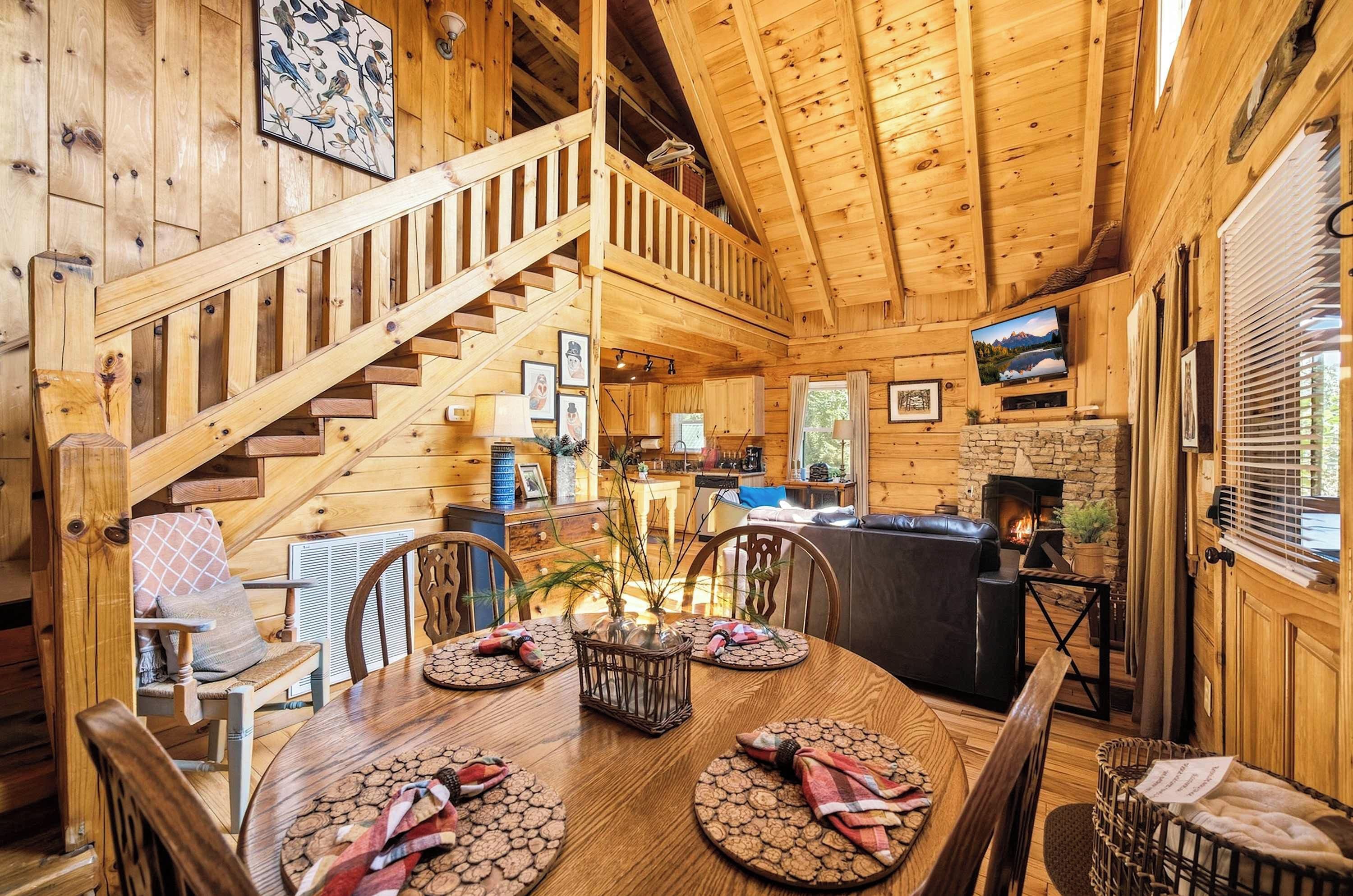 A wooden cabin interior with a dining area, kitchen, and a staircase leading to an upper level.