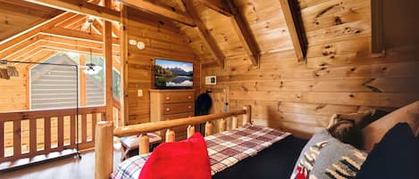 Interior acogedor de una cabaña con cama, televisor montado en la pared y una terraza de madera afuera.