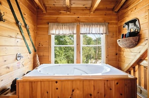 A wooden cabin with a large wooden bathtub, a window with curtains, and a basket on the wall.