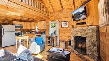 L’intérieur d’une cabane en rondins présente une cheminée en pierre, des murs en bois, un coin cuisine et un espace salon avec un téléviseur fixé au mur.