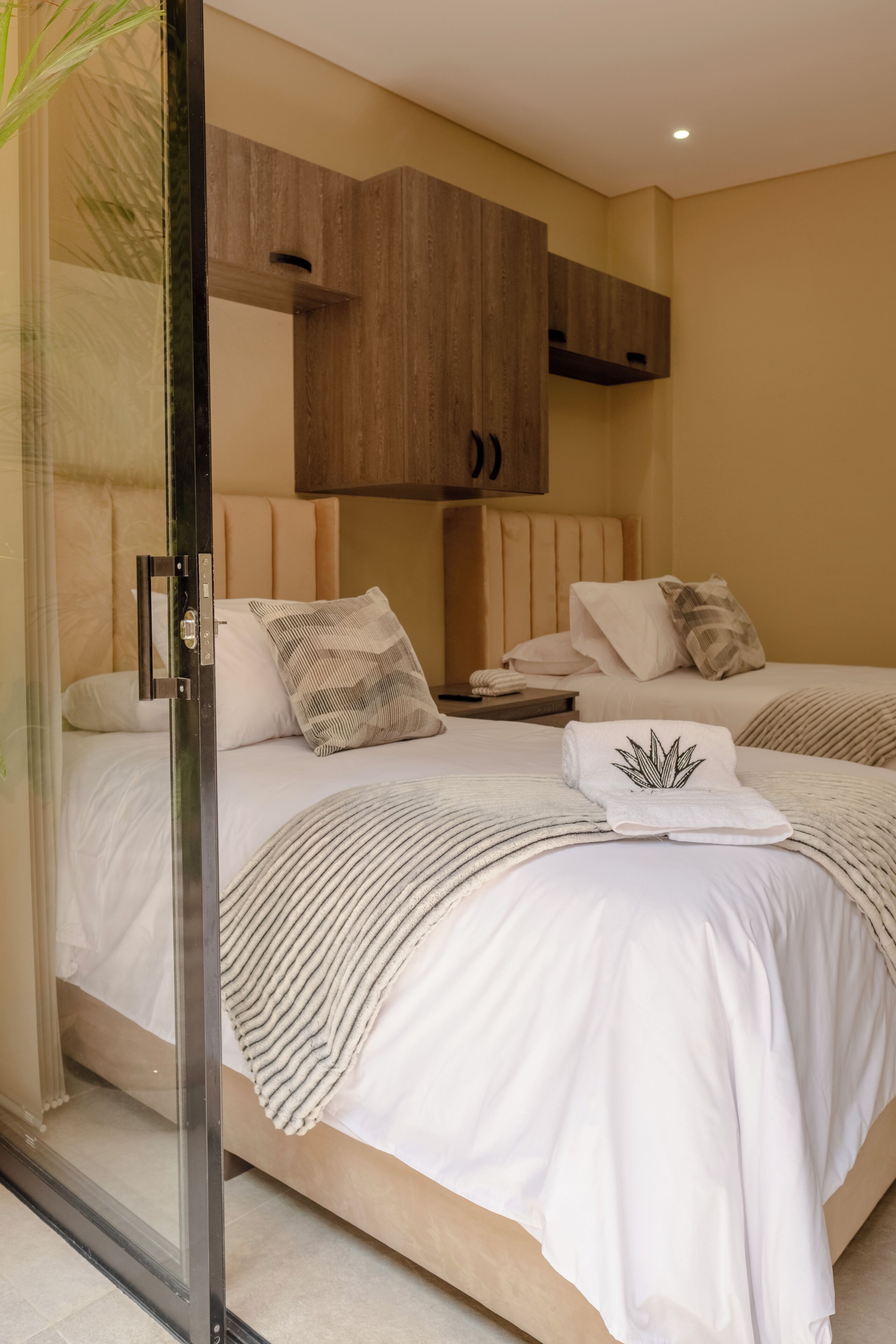 A hotel room with two beds, a wooden headboard, a glass door, and a small bedside table.