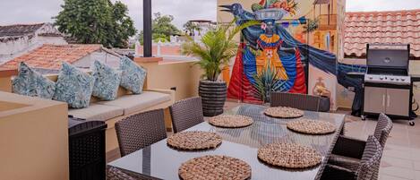 Eine Terrasse im Freien mit einem Esstisch, Korbmöbeln, einem Grill und einem Wandgemälde.