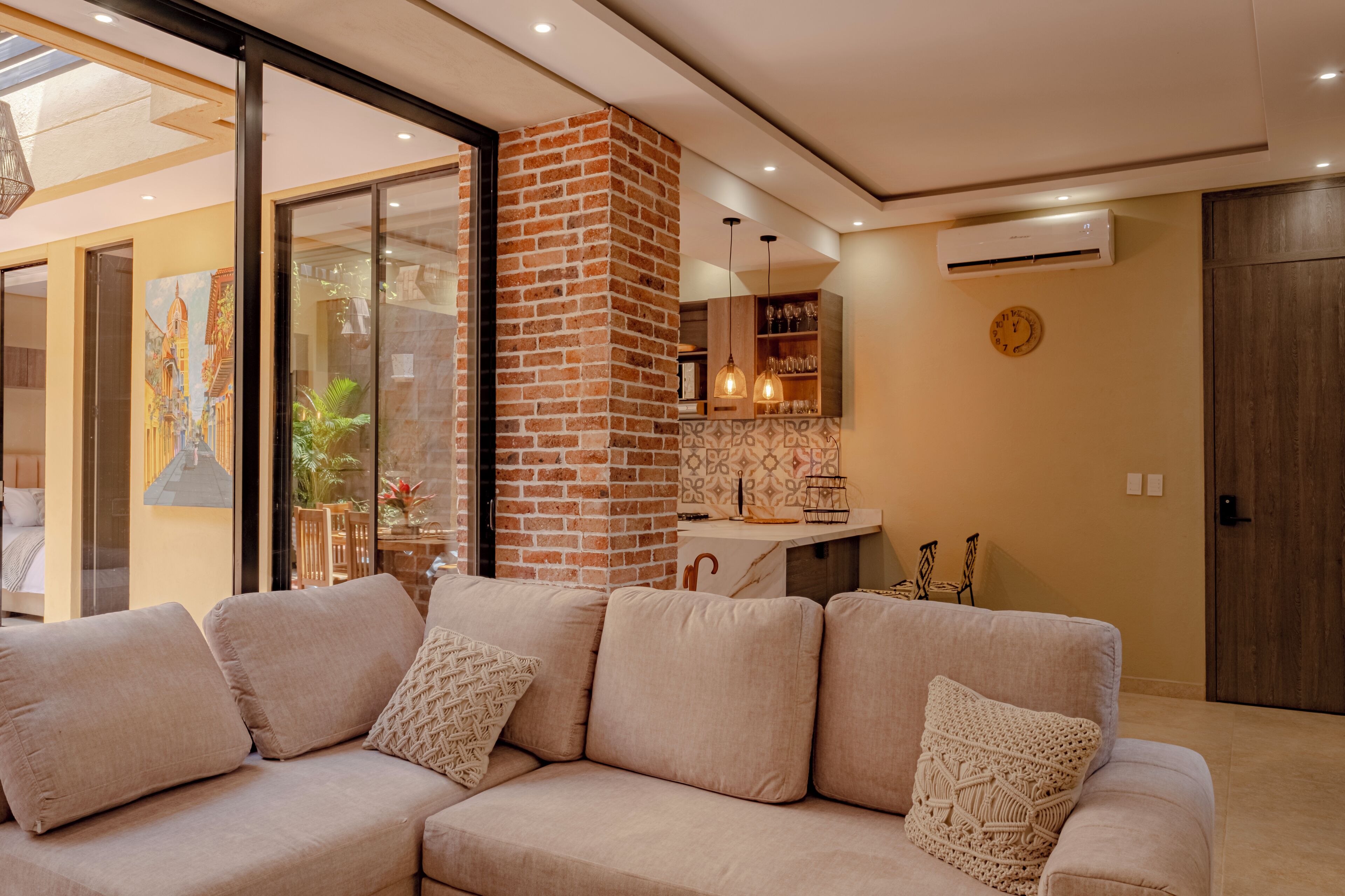 Un salon moderne avec un canapé beige, un mur d’accent en briques et une porte vitrée donnant sur un balcon.