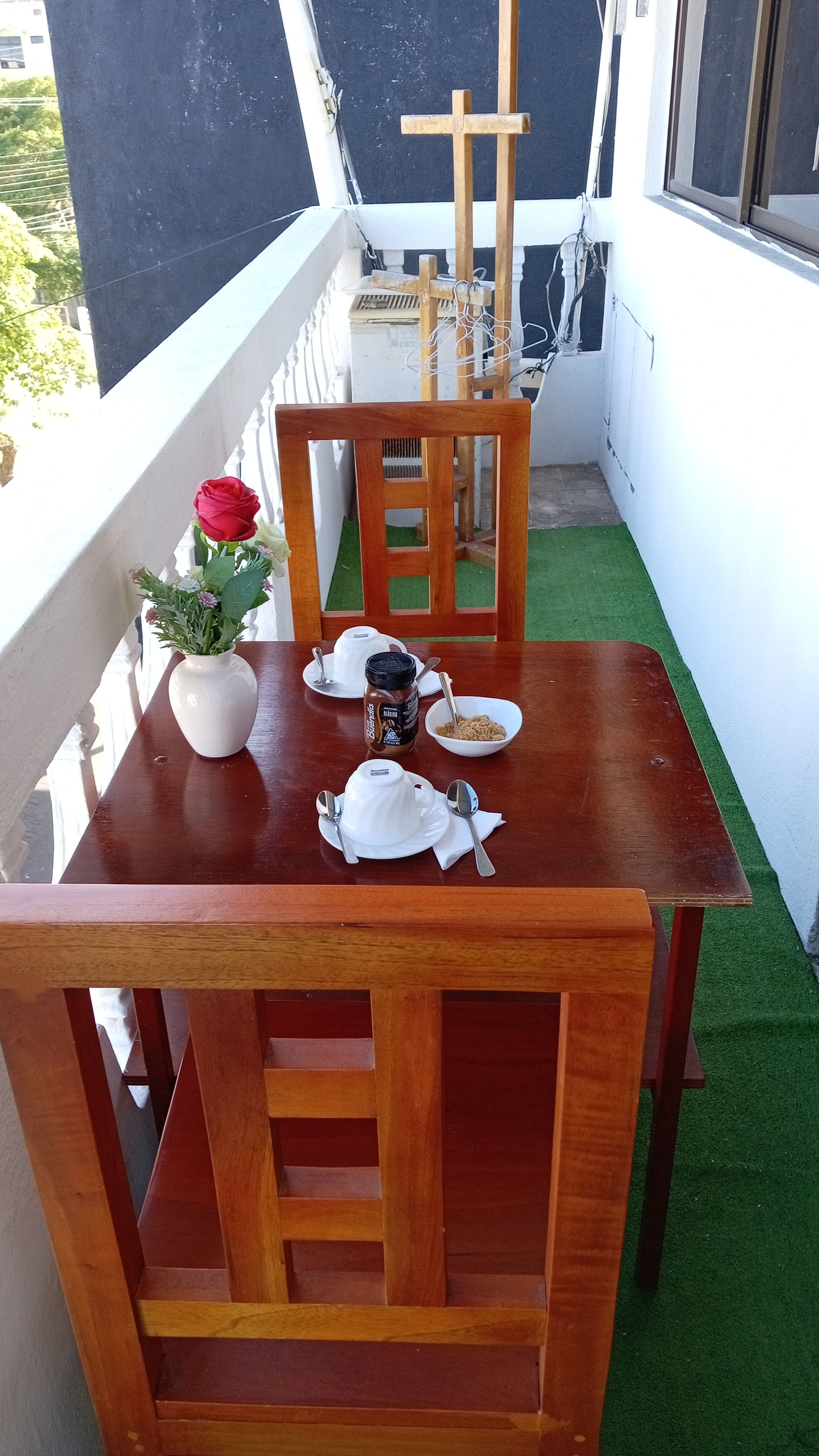 Ein Holztisch mit einer weißen Vase, einem Erdnussbutterglas, einer Schüssel Müsli und einer roten Rose auf einem Balkon mit einem weißen Geländer und Blick auf Grünflächen.