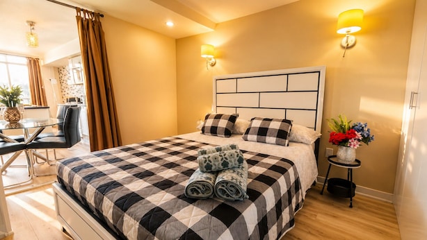A neatly made bed with a checkered bedspread, two wall-mounted lamps, a small table with a vase of flowers, and a dining area with chairs and a table.