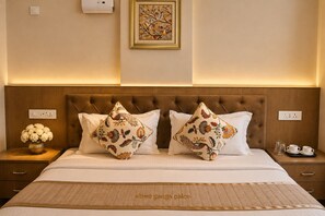 Quarto de hotel com cama, duas almofadas decorativas, mesa de cabeceira com um vaso de flores e uma obra de arte emoldurada na parede.