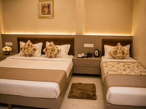 A hotel room with two beds, each with floral-patterned pillows, a nightstand with a phone and a kettle, and a vase with flowers on the left side. - HOTEL SHREE GANGA PALACE (Gwalior)