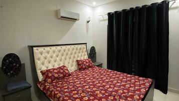 A bedroom with a bed, a nightstand, an air conditioning unit, a ceiling fan, and a black curtain.