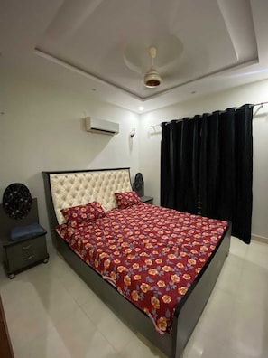 A bedroom with a bed, a nightstand, an air conditioning unit, a ceiling fan, and a black curtain. - Charming 3 Bedroom Bungalow in Karachi with AC & Comfort (Karachi)