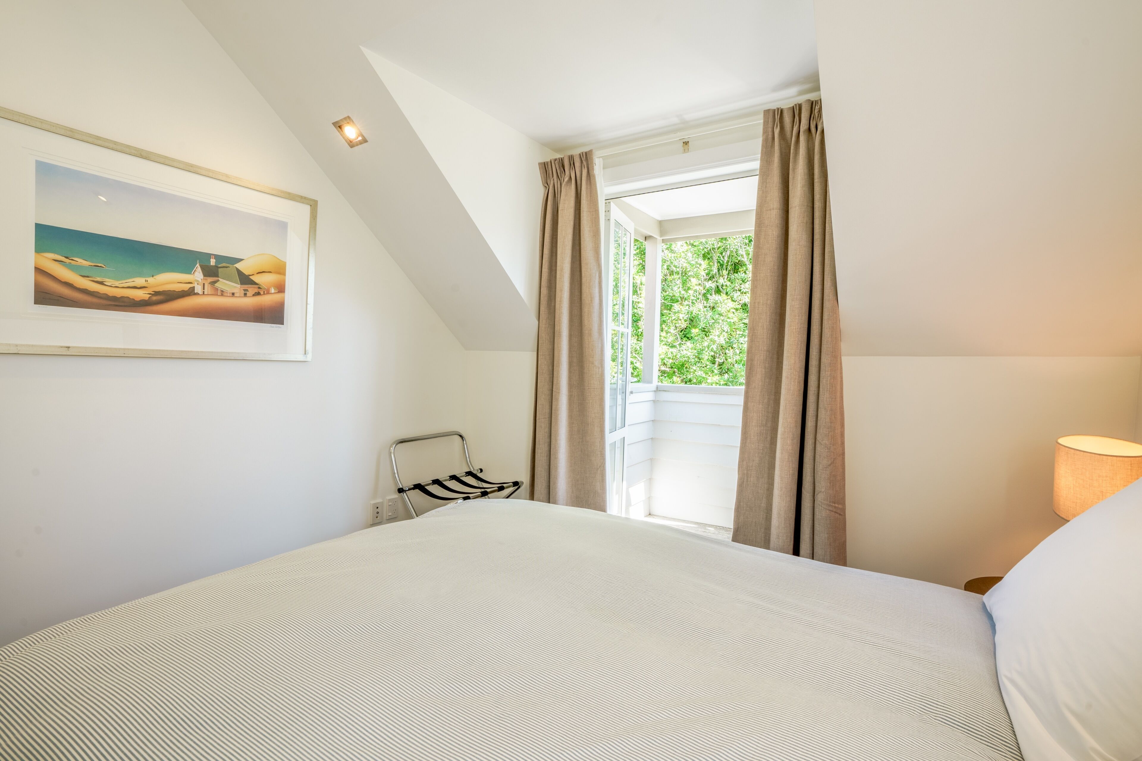 A bedroom with a bed, a painting on the wall, a window with curtains, and a lamp.