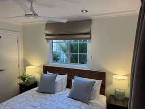 A bedroom with a bed, two bedside lamps, a ceiling fan, a window with a view of greenery, and a door. - Crab Shack Cottage: 4BR in Russell with Pool & Spa, 300m to Waterfront (Russell)